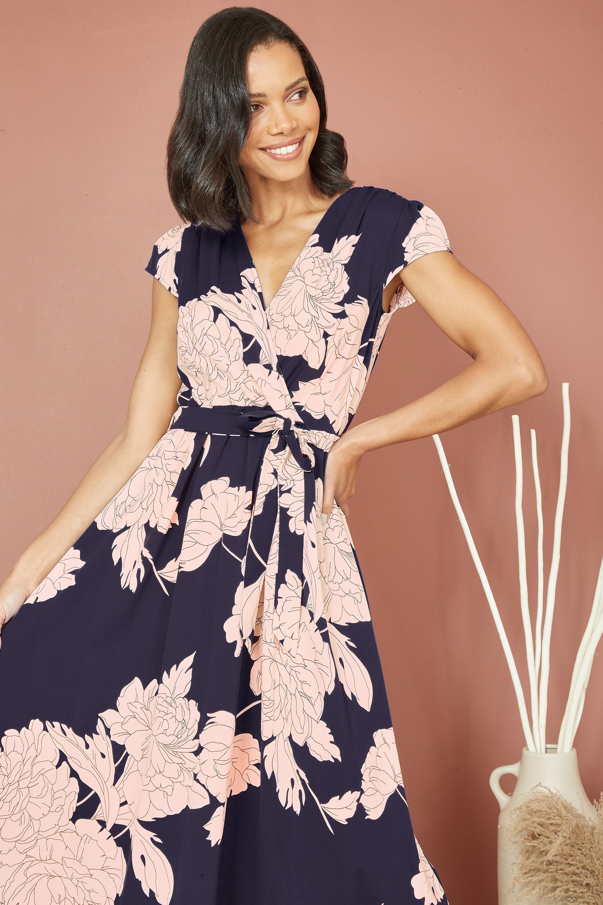 Yumi Navy Blossom Print Wrap Midi With Dipped Hem — front lifestyle view showing navy wrap dress with pink blossom print, frill trim, short sleeves and tie waistline flowing below the knee