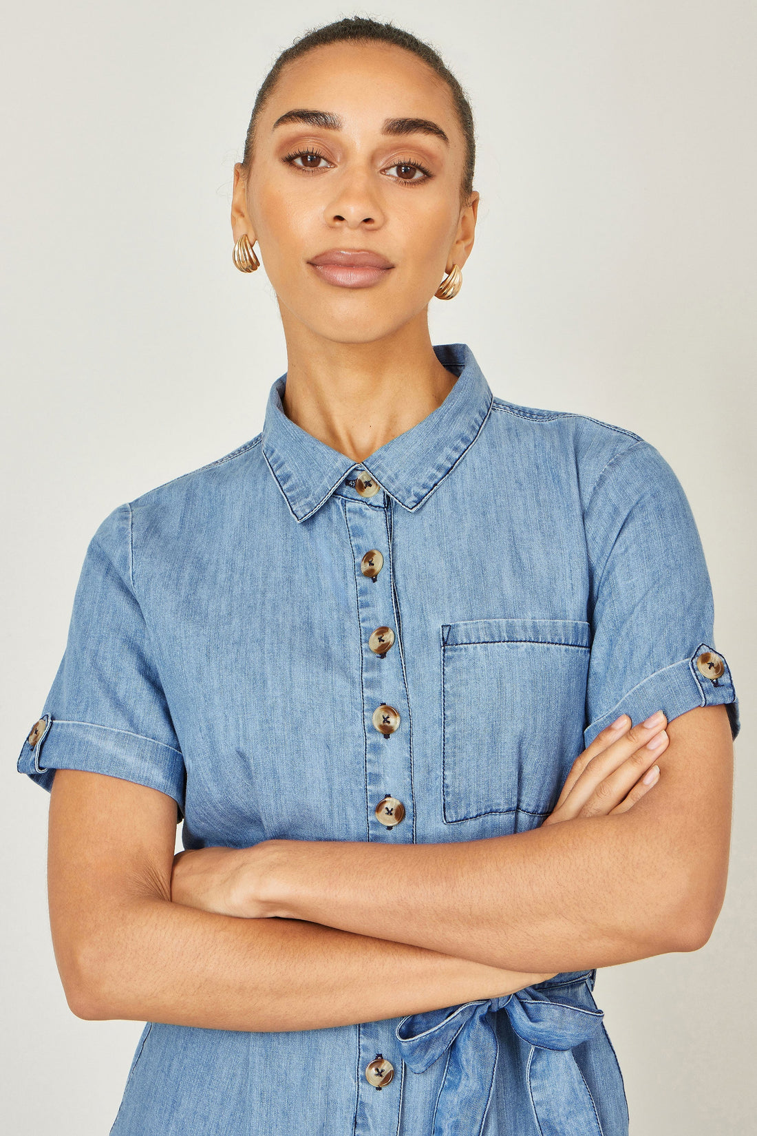 Yumi Blue Chambray Light Denim Button Jumpsuit — front lifestyle view showing 100% cotton chambray jumpsuit with button front, waist tie, side pockets and flared culotte trousers in light blue denim