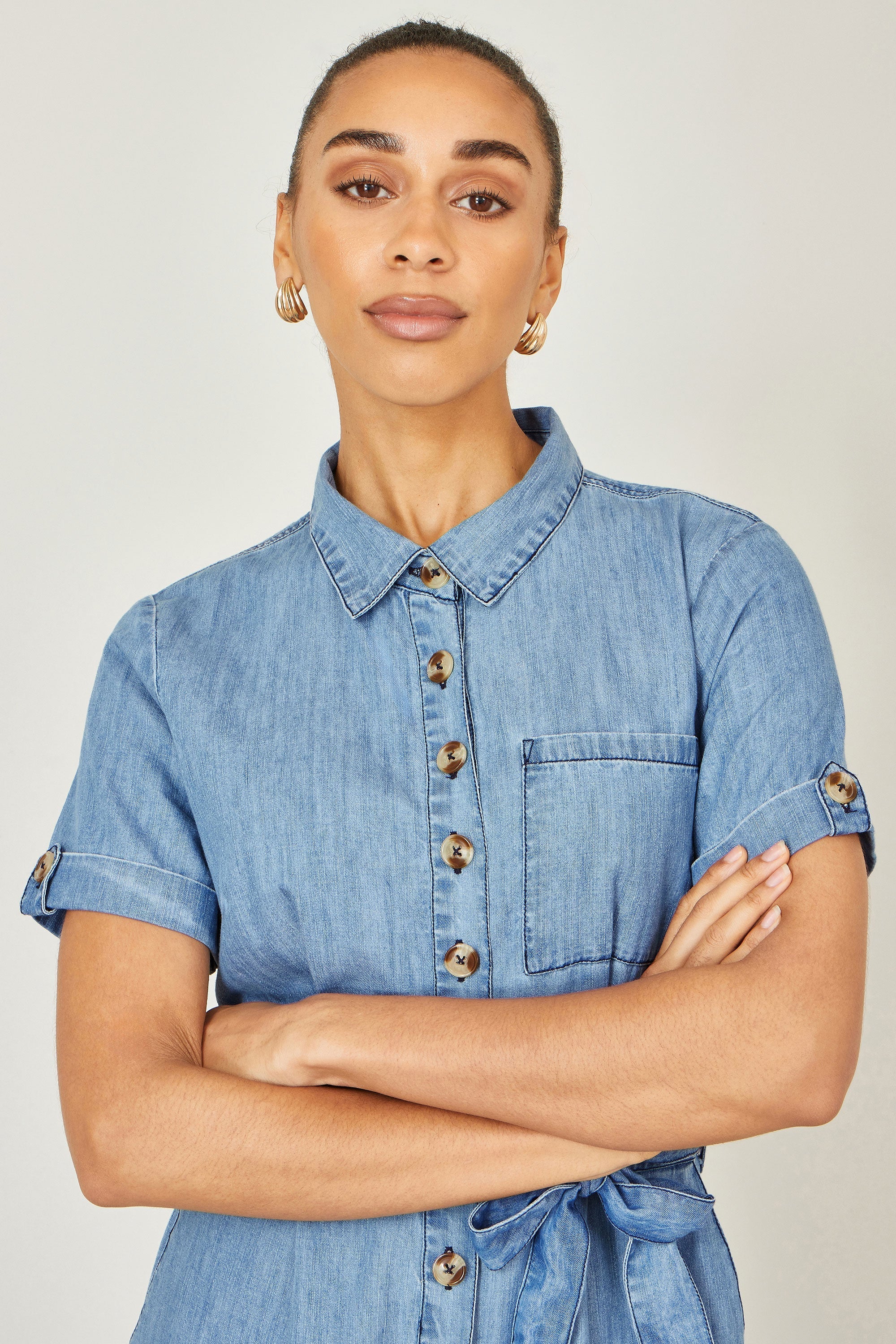 Yumi Blue Chambray Light Denim Button Jumpsuit — front lifestyle view showing 100% cotton chambray jumpsuit with button front, waist tie, side pockets and flared culotte trousers in light blue denim