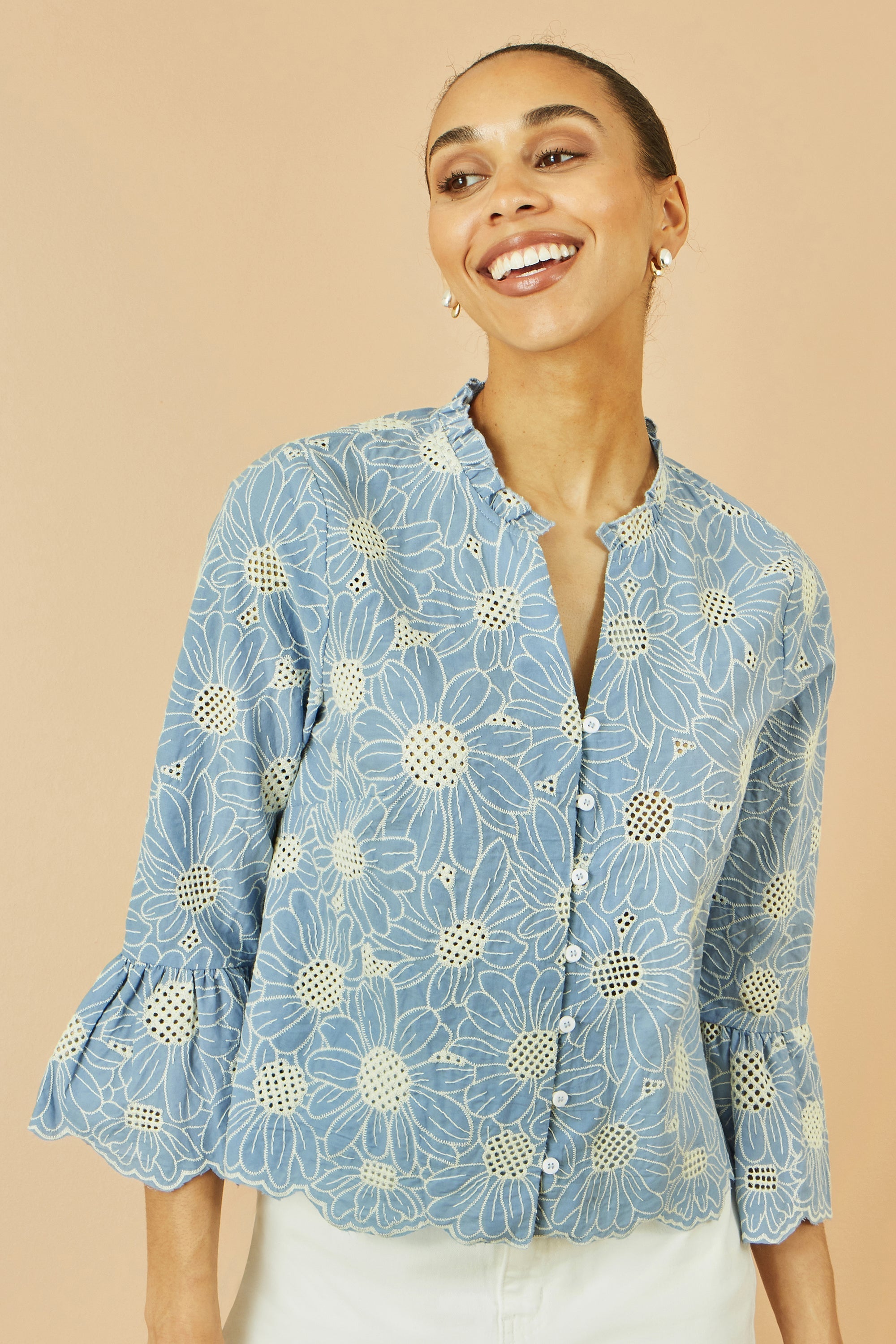 Yumi Blue Daisy Broderie Shirt with fluted sleeves and frill collar shown on a model in a summer setting displaying the daisy broderie anglaise design
