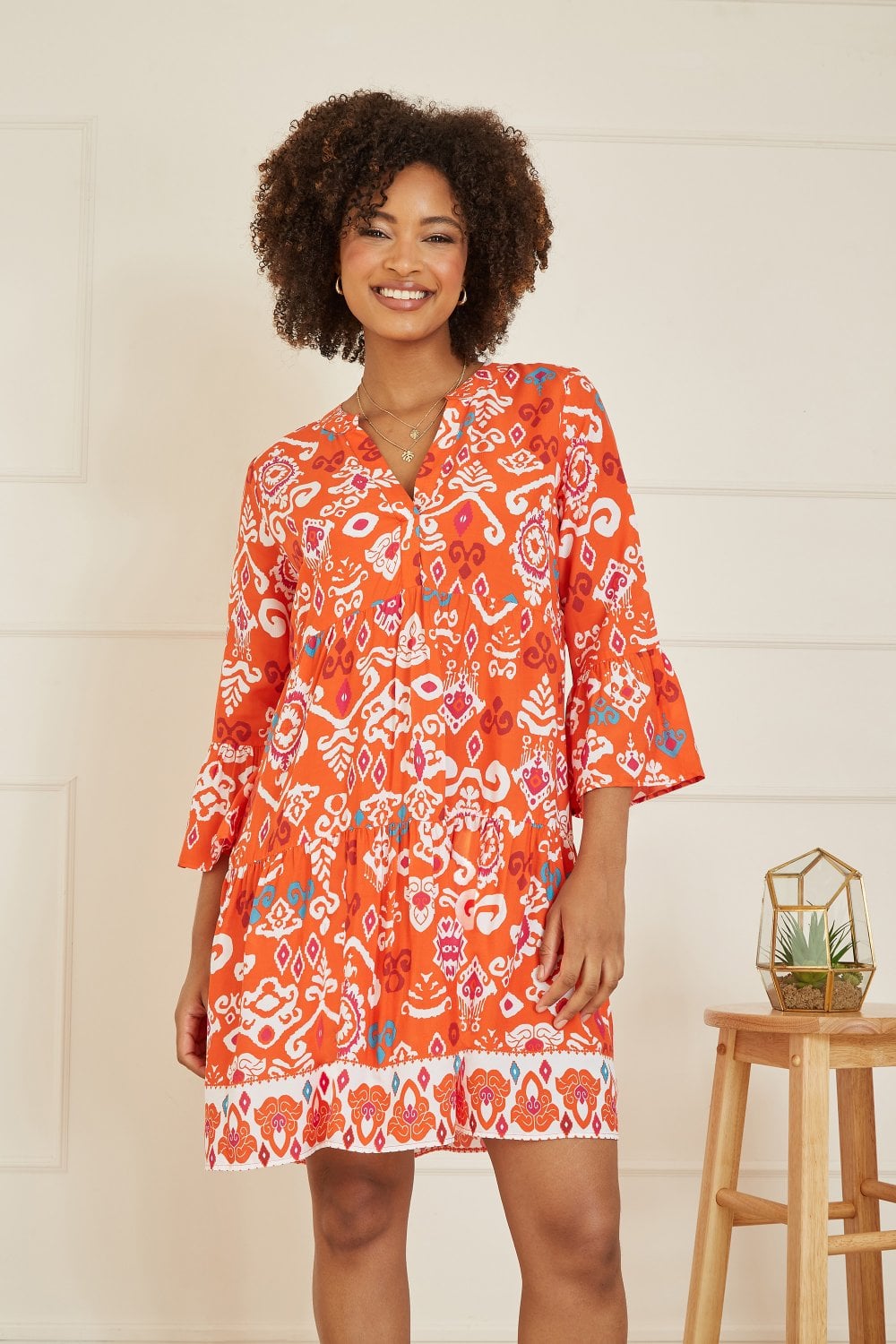 Yumi Orange Ikat Print 3/4 Sleeve Tunic Dress in bold orange pattern with bell flare sleeves and relaxed fit shown on a model in a sunny setting